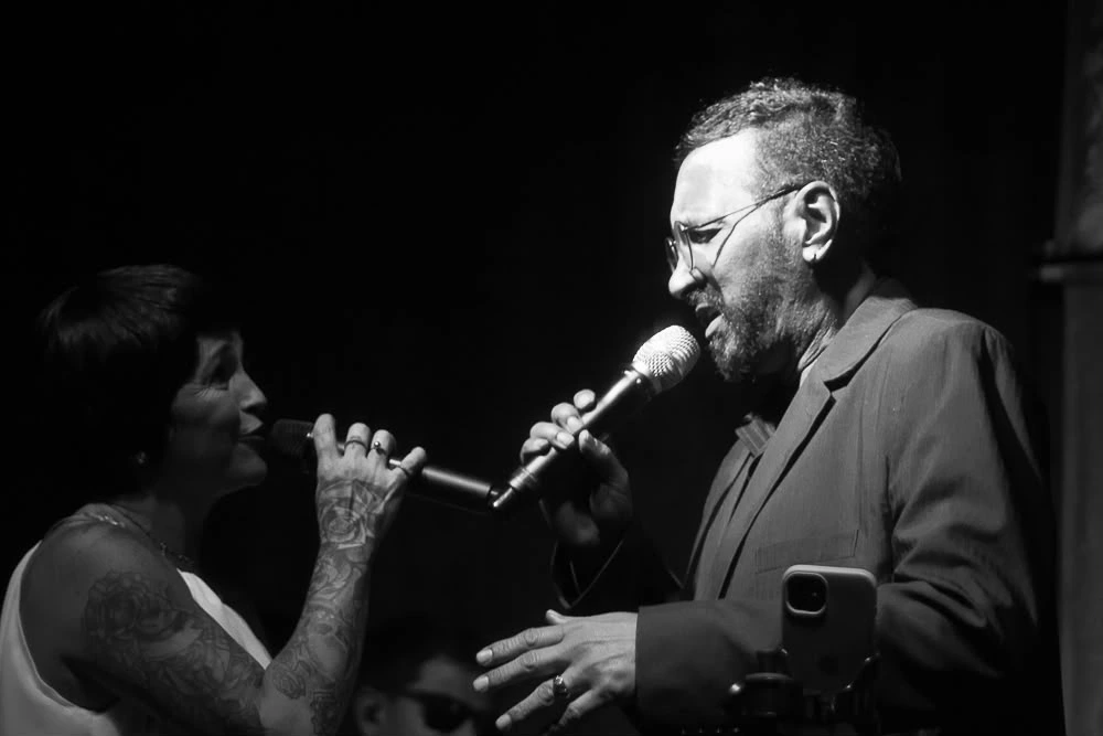 Cantora interpretando clássicos de Elis Regina ao vivo no palco com microfone vintage e iluminação dramática